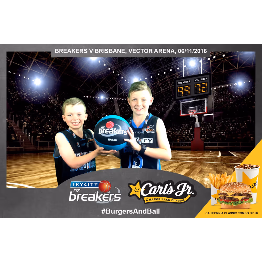 Two boys in basketball jerseys holding an N.Z. Breakers ball on court with scoreboard and Carl's Jr. ad.