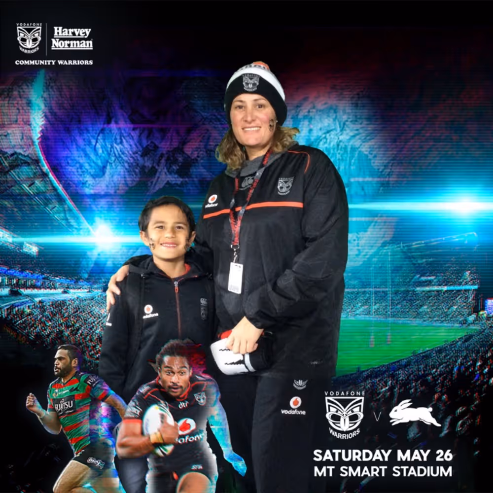 Two people in Vodafone Warriors apparel smiling with stadium and rugby players in the background.