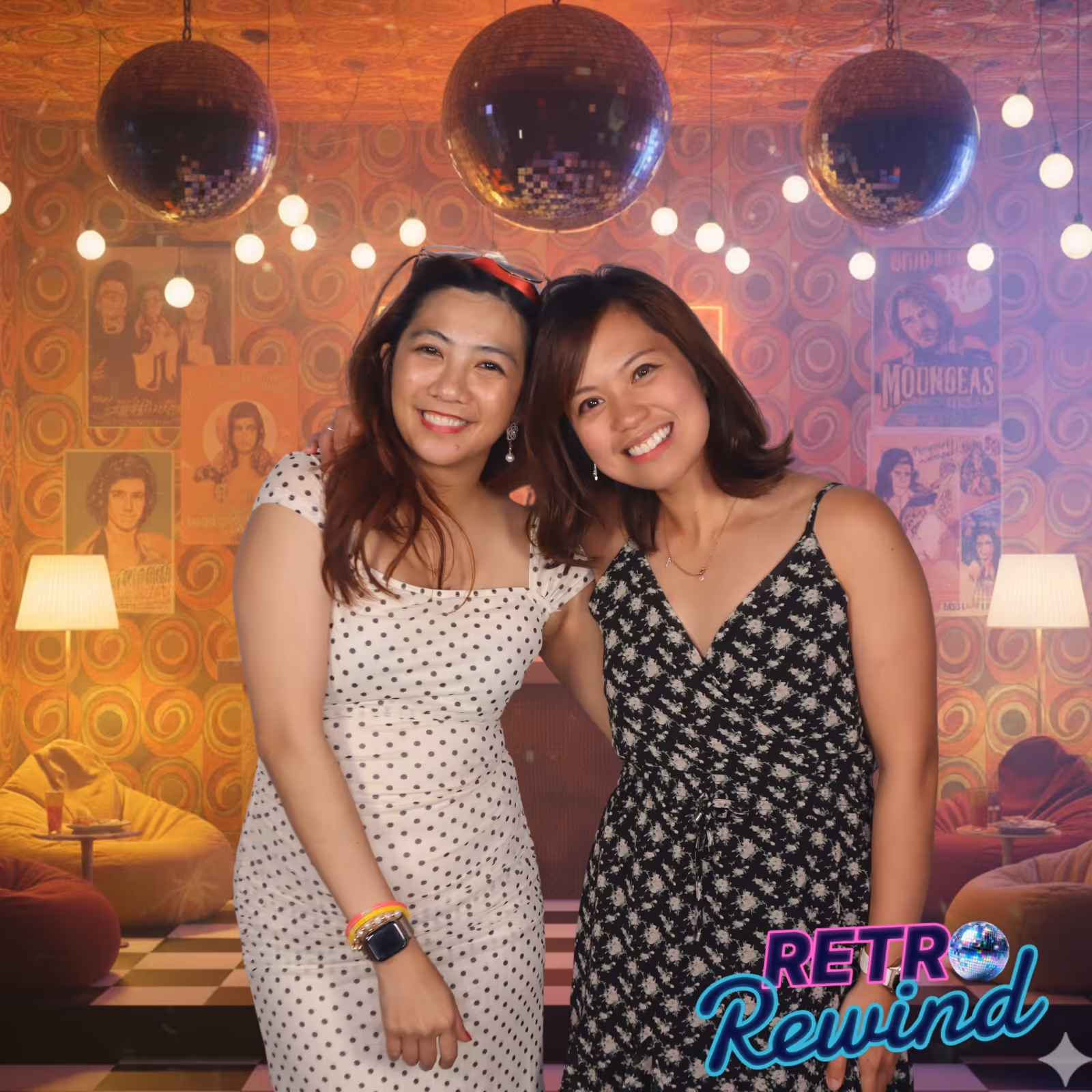 Two women smiling and posing in front of a retro-themed disco background with hanging disco balls.