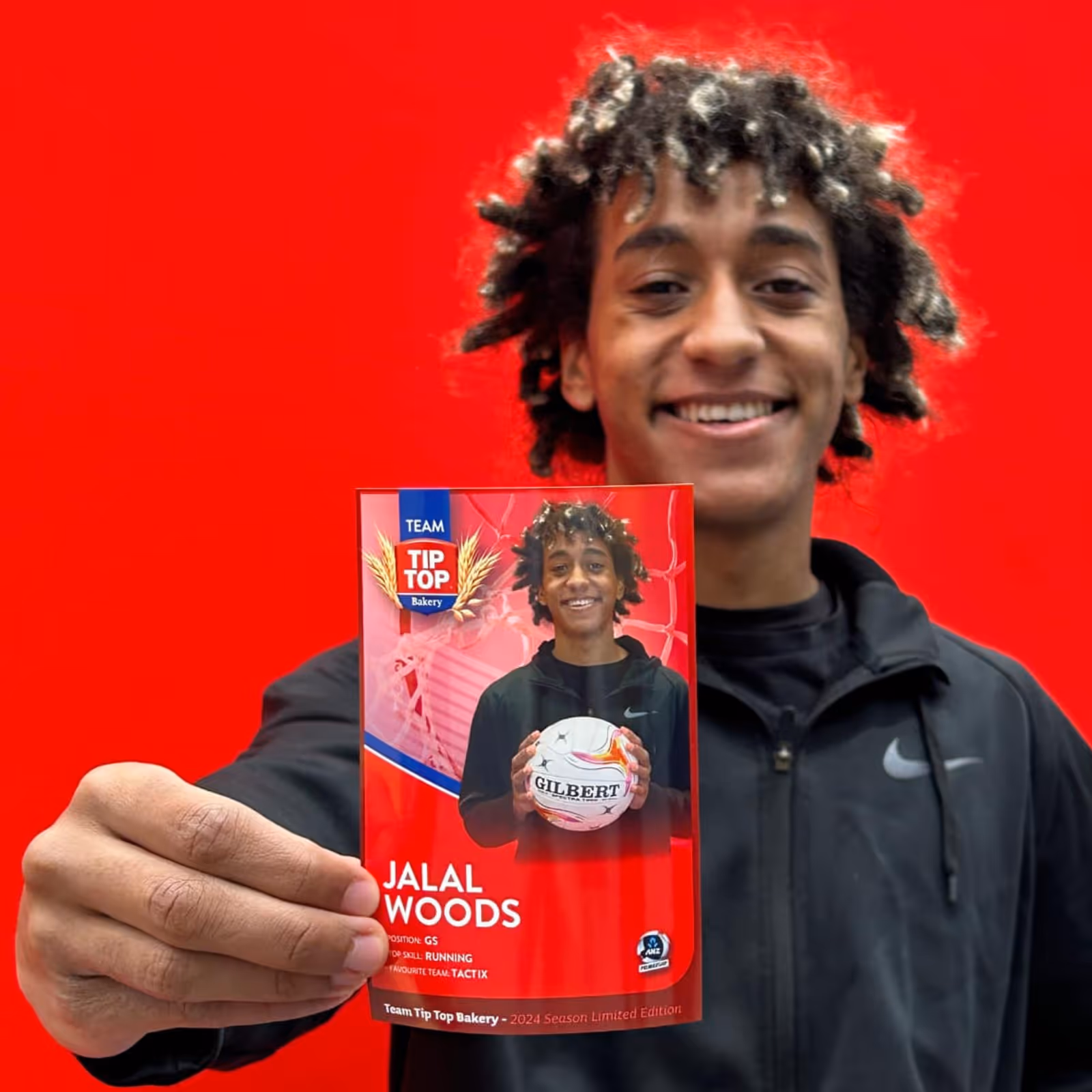 Young man holding a card with his photo, name Jalal Woods, and netball details against red wall.