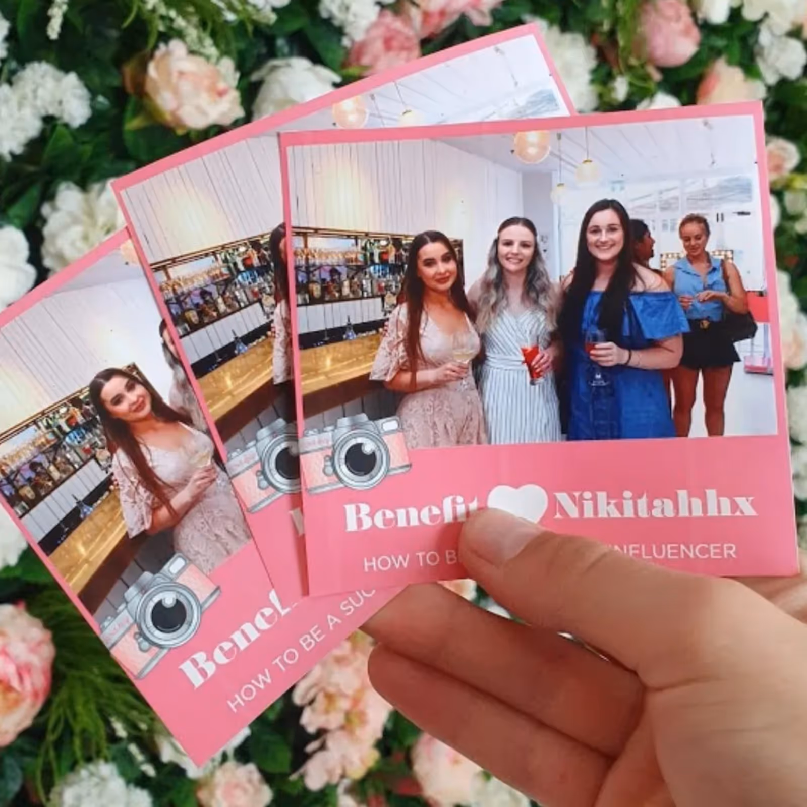 Hand holding pink cards featuring three women holding drinks in a bright room with flowers in background.