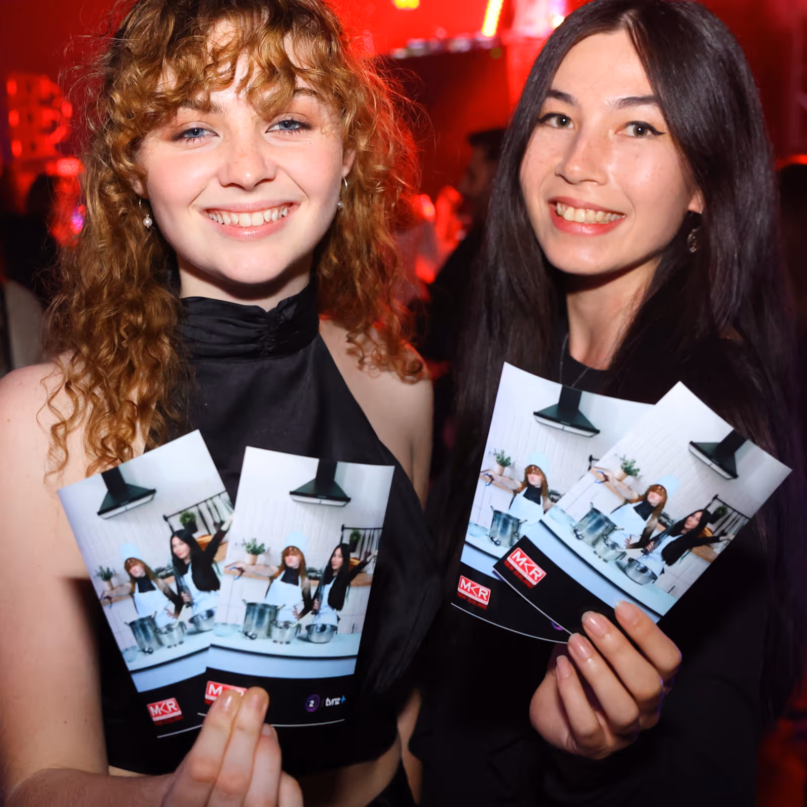 Two smiling women holding flyers with a cooking show image in a dim, red-lit setting.