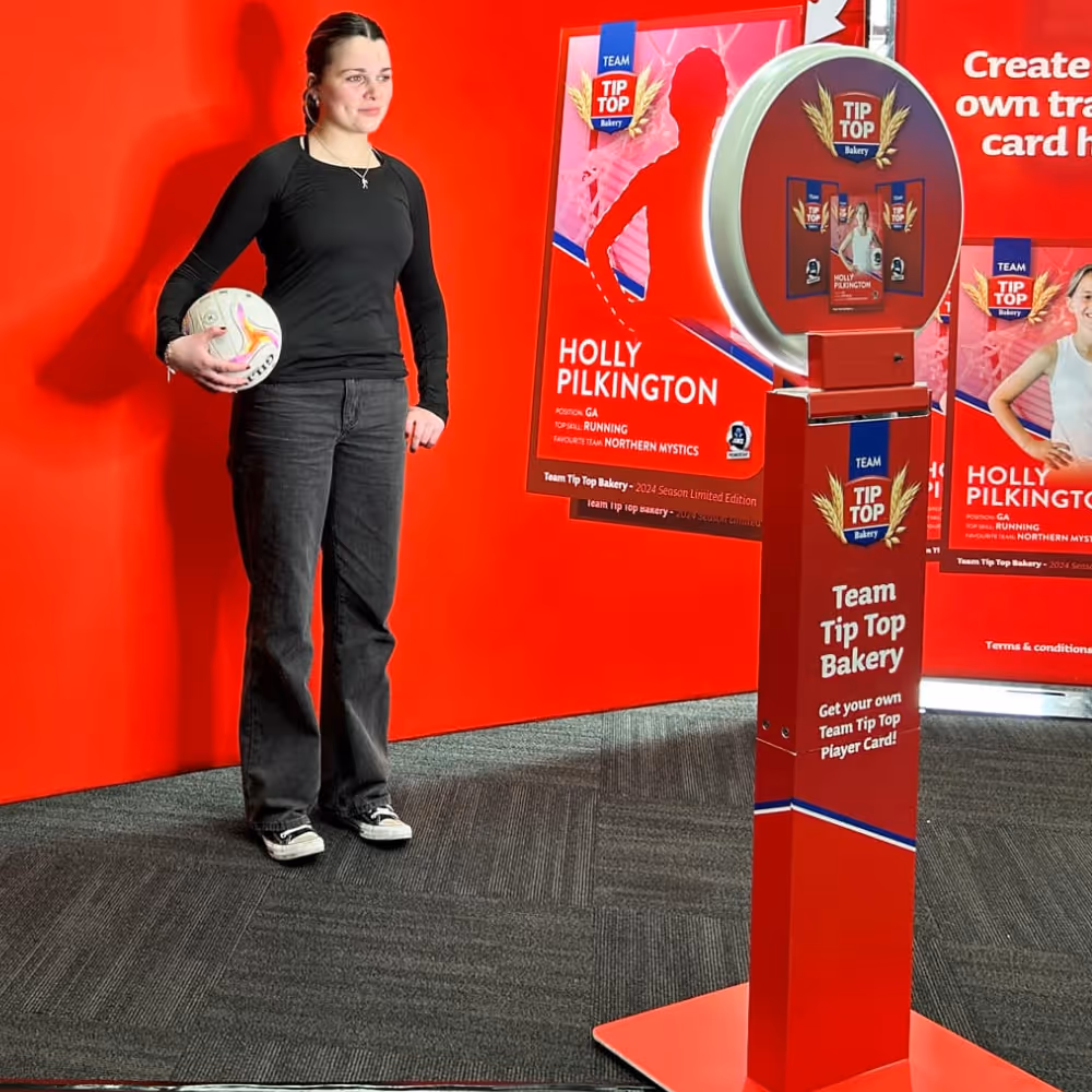 Woman in black holding a netball standing against red wall with Team Tip Top Bakery promotional display.