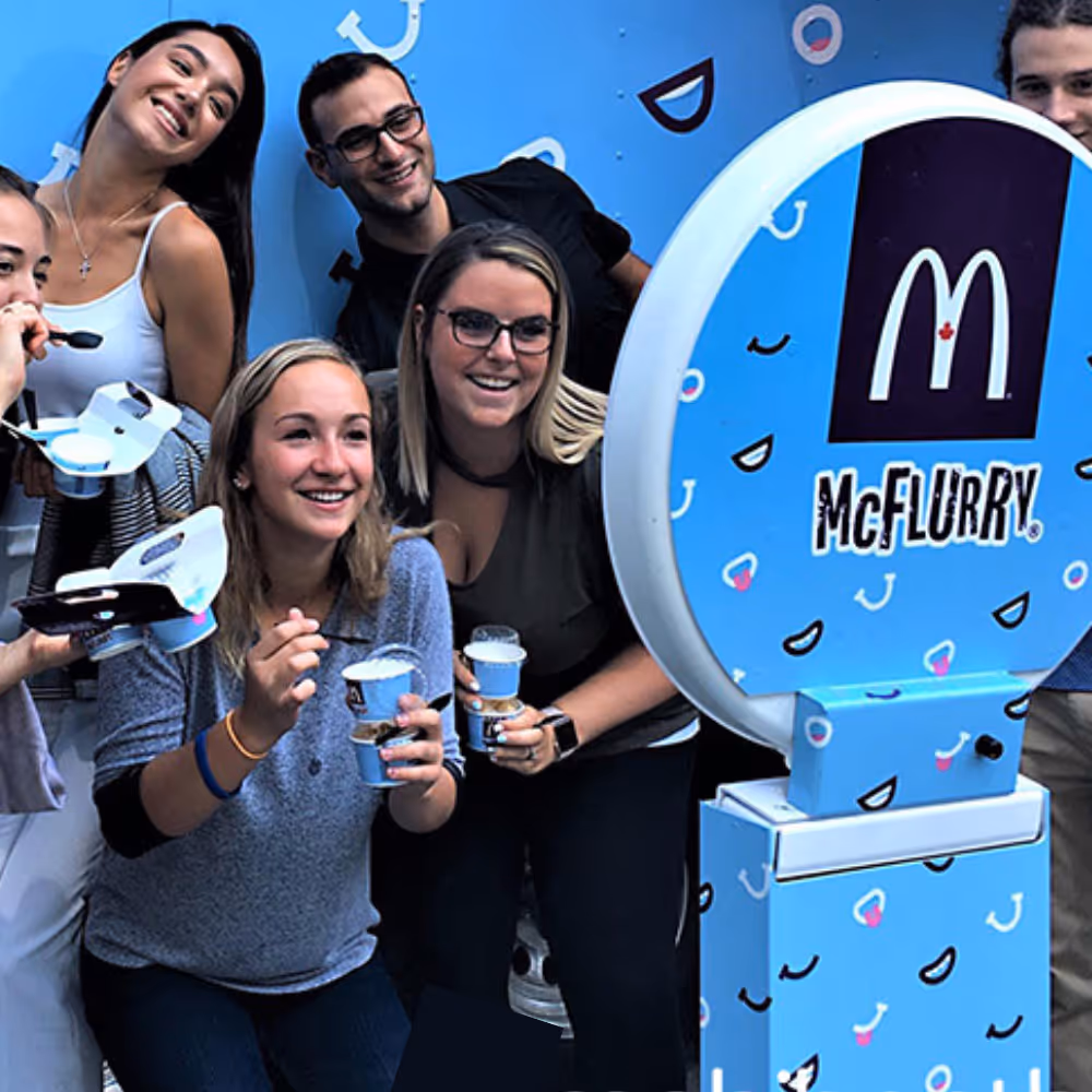 Group of young people happily holding McFlurry ice cream cups near a blue McFlurry sign.