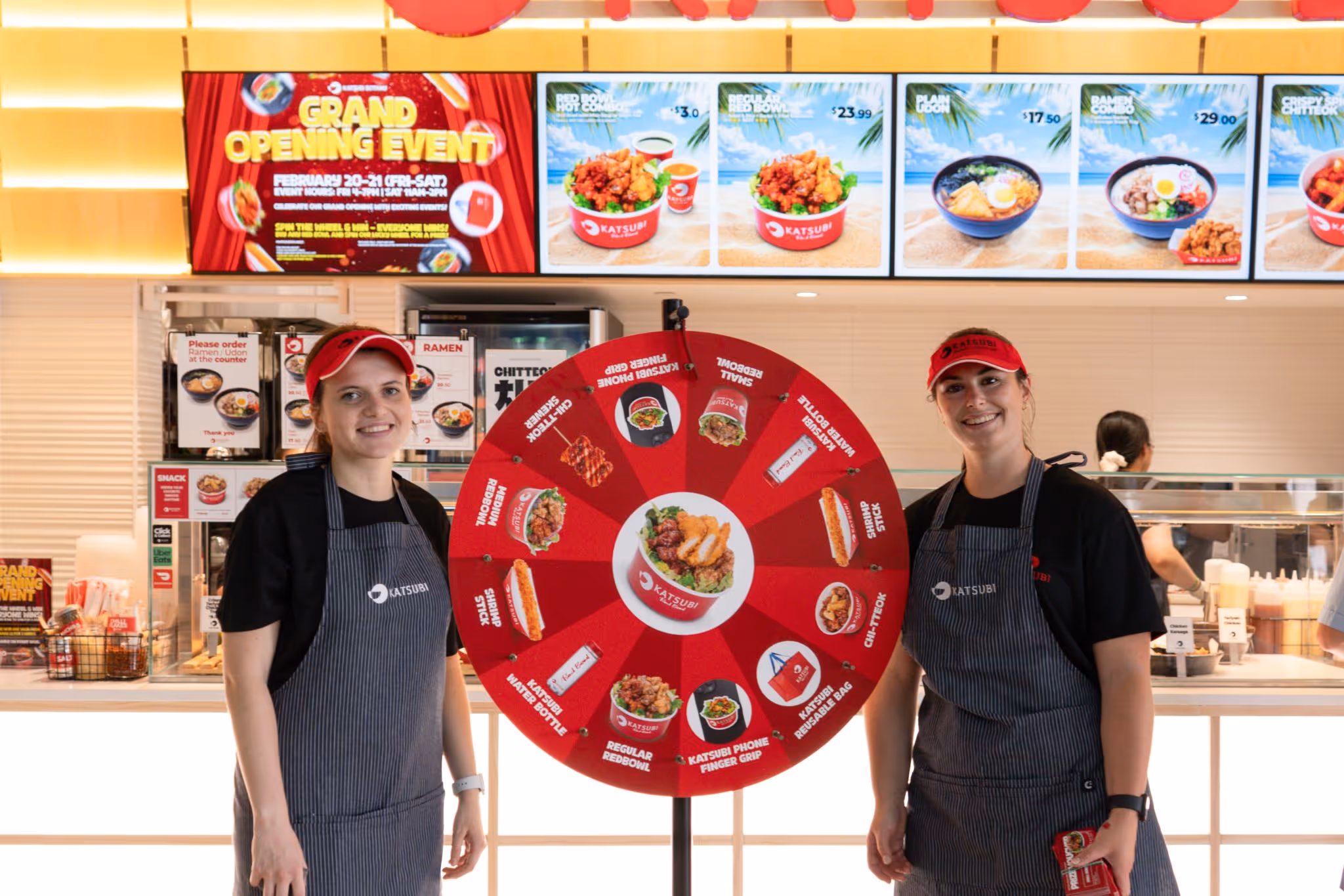 Two smiling staff in aprons and red caps stand beside a red prize wheel in a Japanese food restaurant.