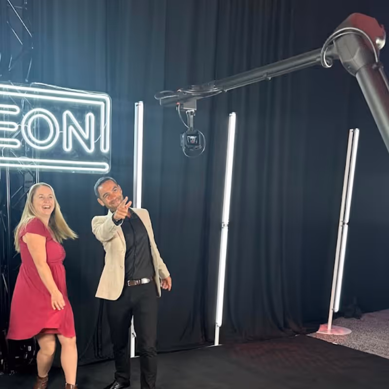 A man and woman smiling and pointing toward a camera in a studio with neon and vertical lights.