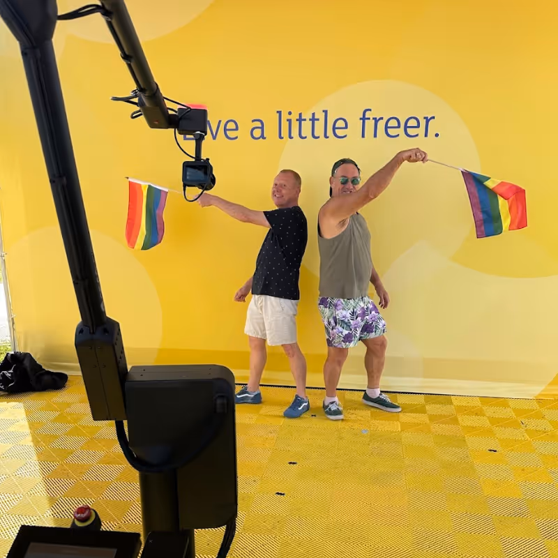 Two men holding rainbow pride flags back to back against a yellow wall with partial text visible.