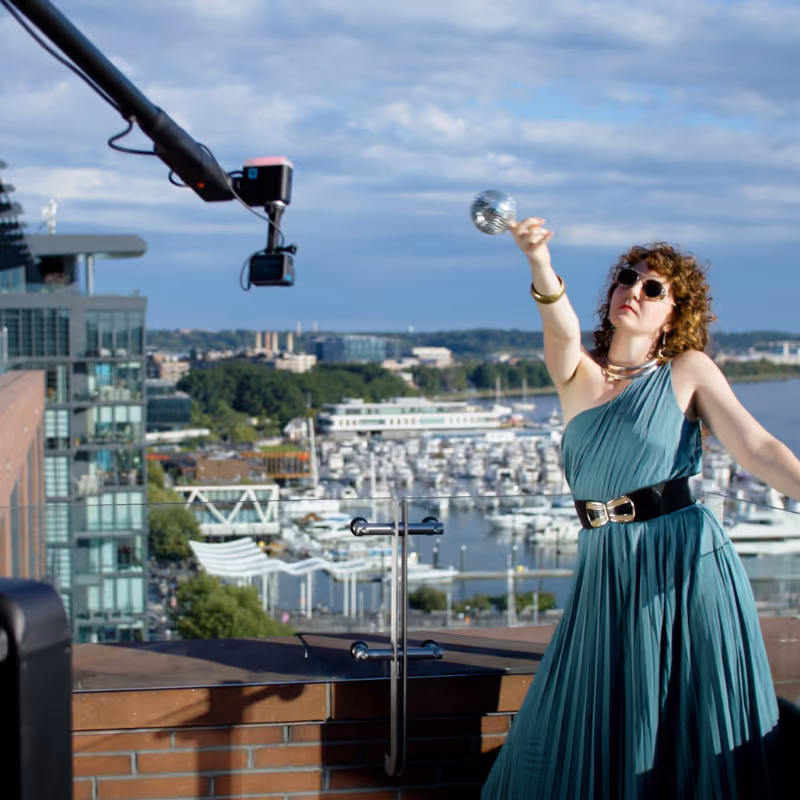 Woman in green dress with sunglasses poses on rooftop with marina and boats in background.