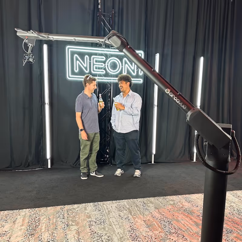 Two men holding drinks and talking in front of a neon sign that reads NEON and black curtains.