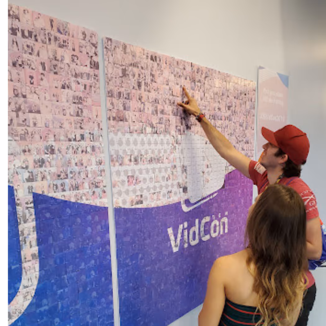 Two people look at and point to a large VidCon mosaic wall made of small individual photos.