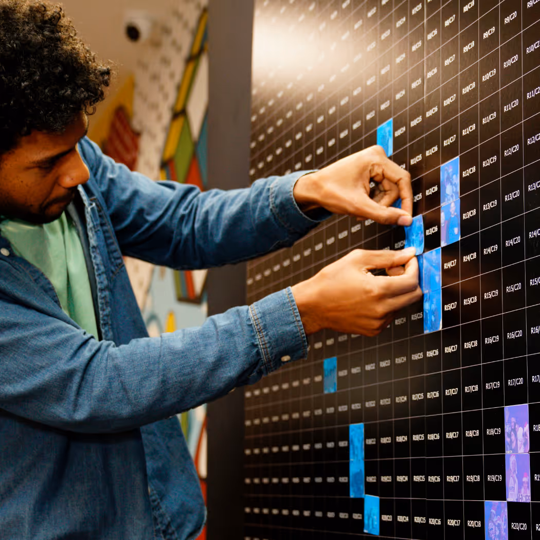 Man placing blue photo squares on a large black grid wall with labeled cells.