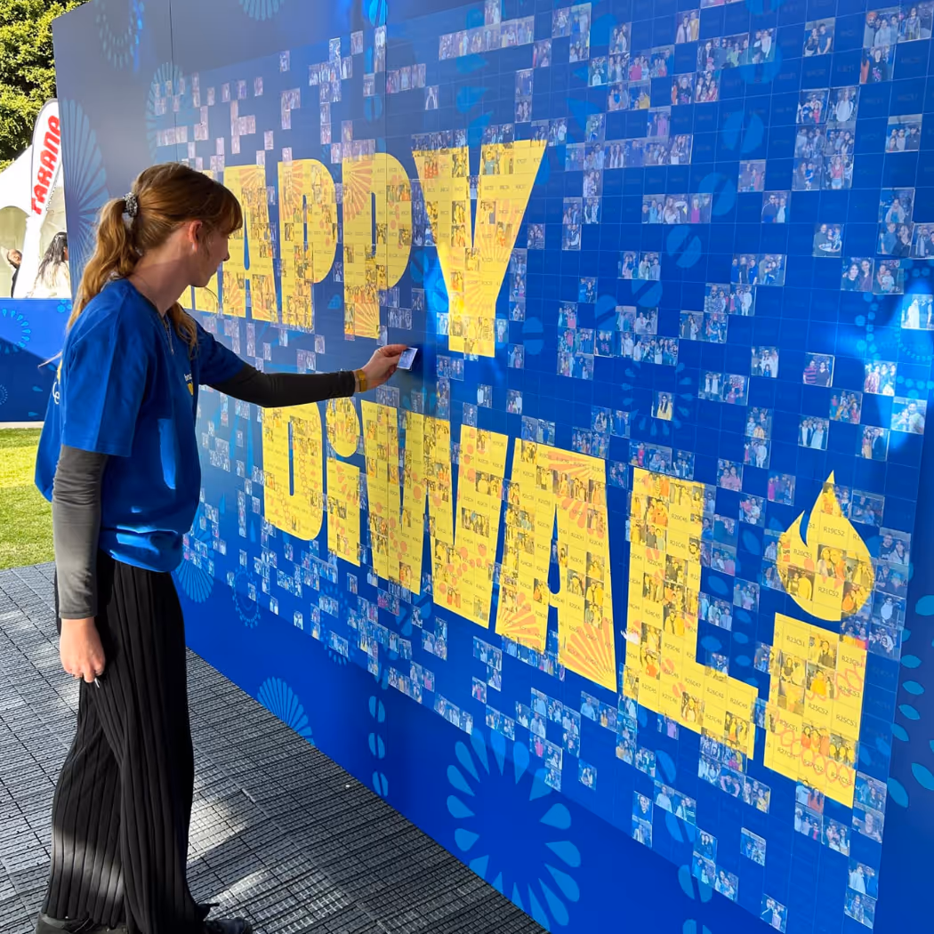 Woman placing a small photo on a large mosaic wall spelling HAPPY DIWALI in yellow letters.