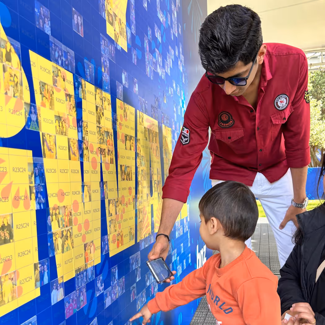 Young man shows a smartphone screen to a child pointing at a colorful photo collage wall.