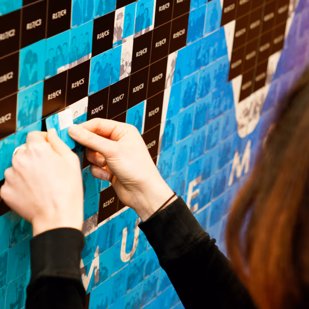 Person placing a small photo tile on a mosaic wall spelling 'MUSEUM' with many blue and black photo tiles.