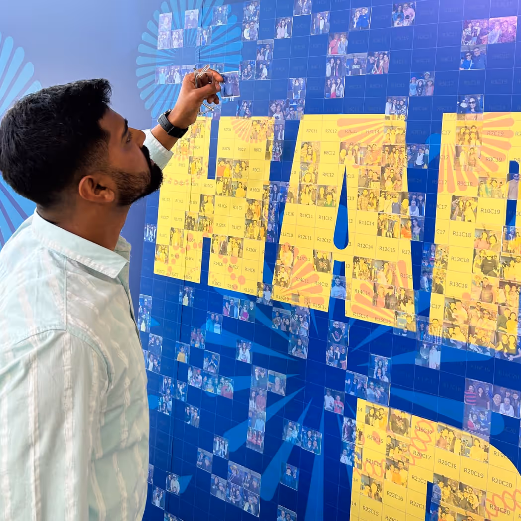 Man holding a photo while looking at a large mosaic wall with yellow text on a blue background.