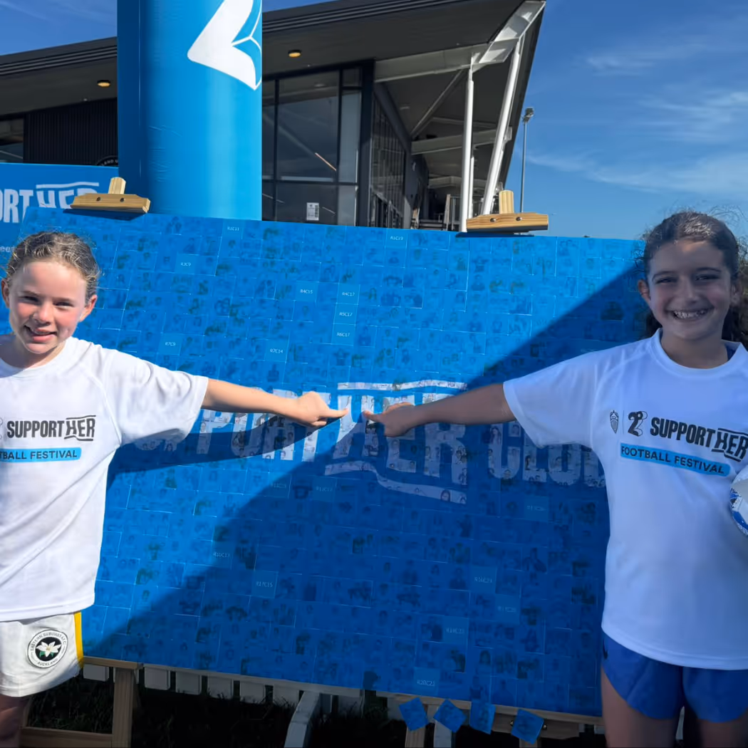 Two girls in white shirts pointing at a blue board with 'SUPPORT HER' text outdoors.