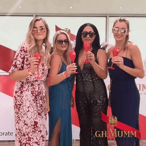 Four women wearing sunglasses in dresses holding red champagne glasses posing at a GH Mumm branded event.