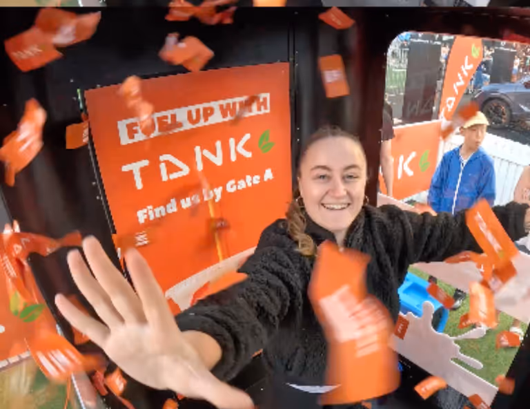 Smiling woman reaches out as orange prize cards fall inside a machine with Tank brand ads.