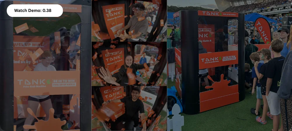 Outdoor event with people playing an orange Tank prize grab machine and others watching in line.