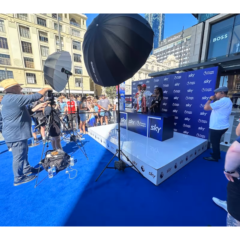 Outdoor photo shoot showing people near a Sky Premier League stage with trophies and lighting equipment.
