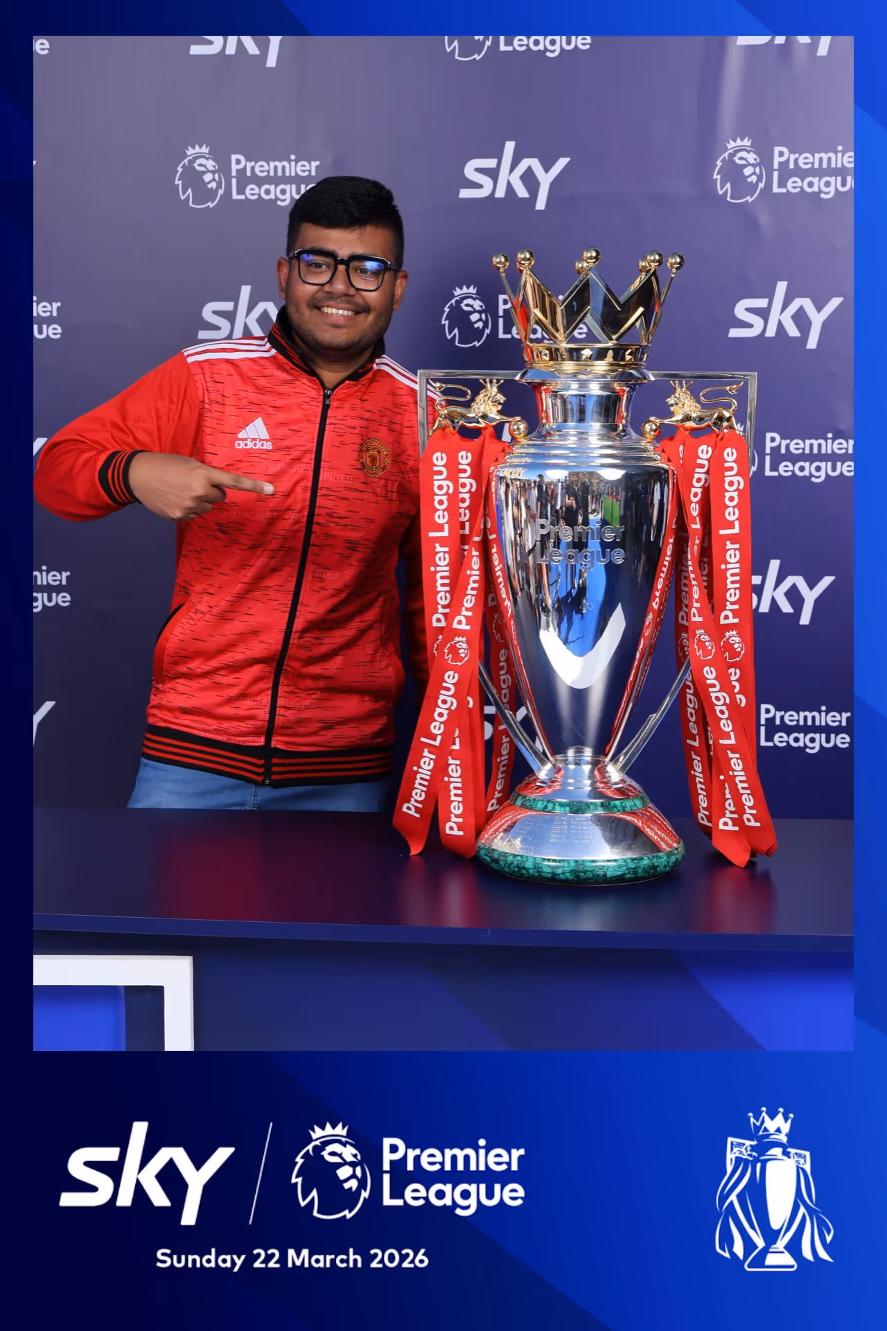 Man in red Manchester United jacket pointing at the Premier League trophy with red ribbons.