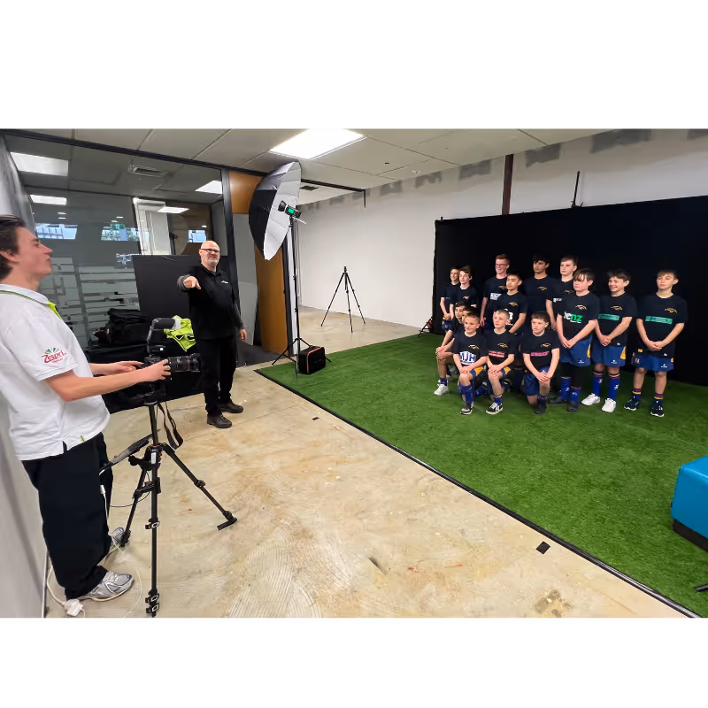 Children's sports team posing on artificial turf for a group photo with photographers and lighting set up.