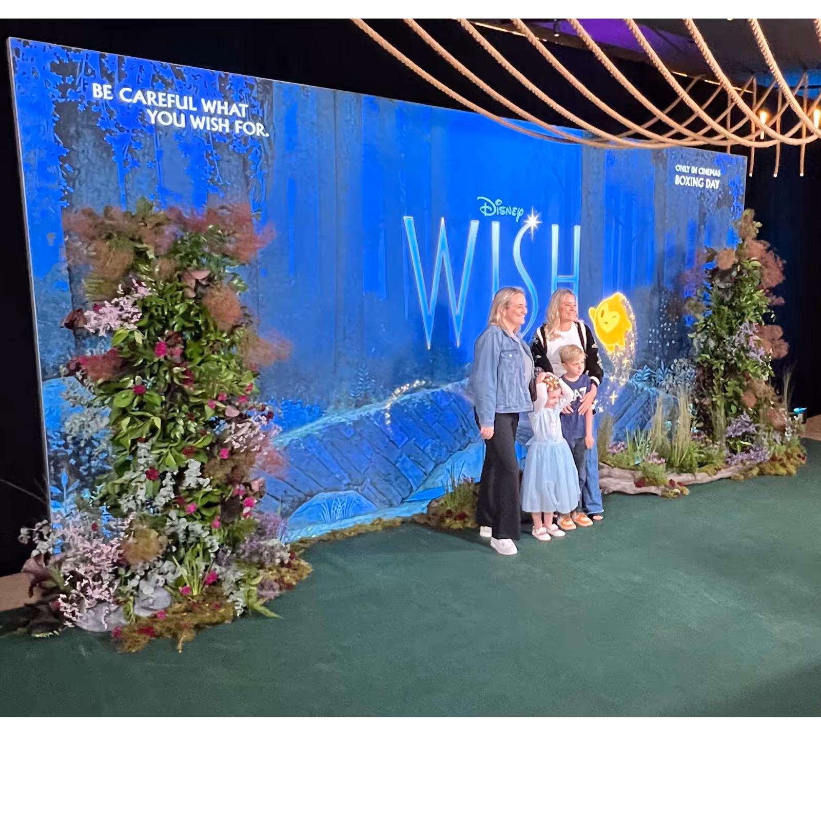 Four people posing in front of a Disney Wish movie backdrop decorated with flowers and greenery.