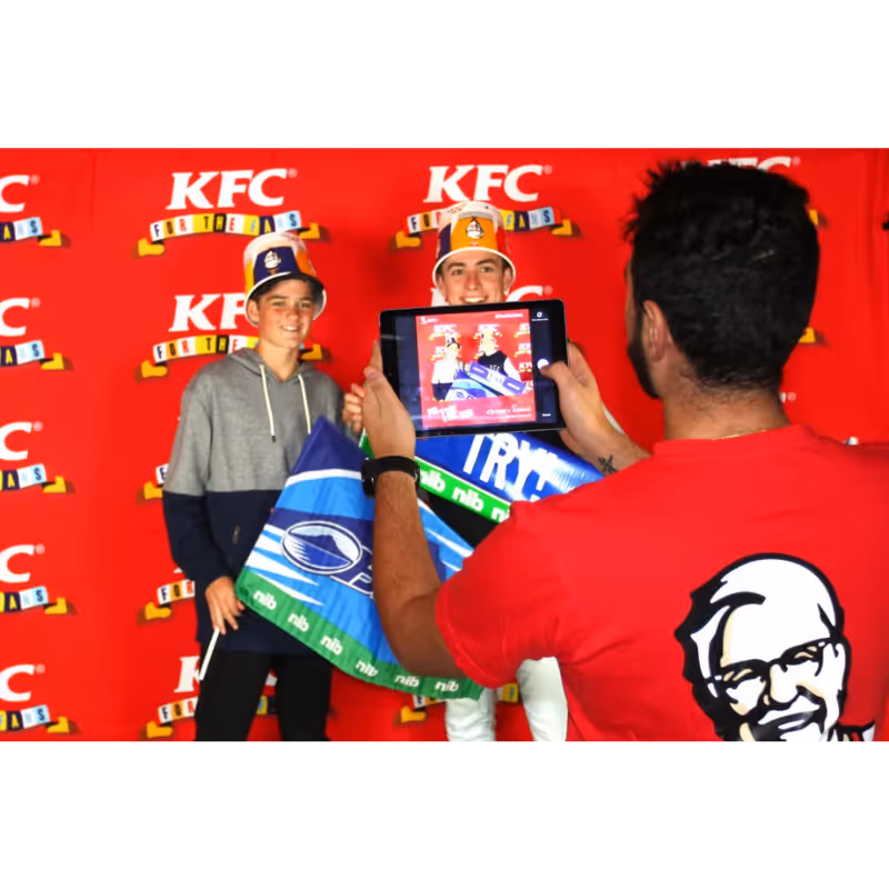 Two boys wearing KFC buckets pose for a photo taken by a man with a tablet in front of a KFC backdrop.