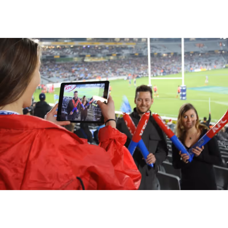 A woman in red takes a photo with a tablet of two people at a sports stadium holding red and blue KFC foam sticks.