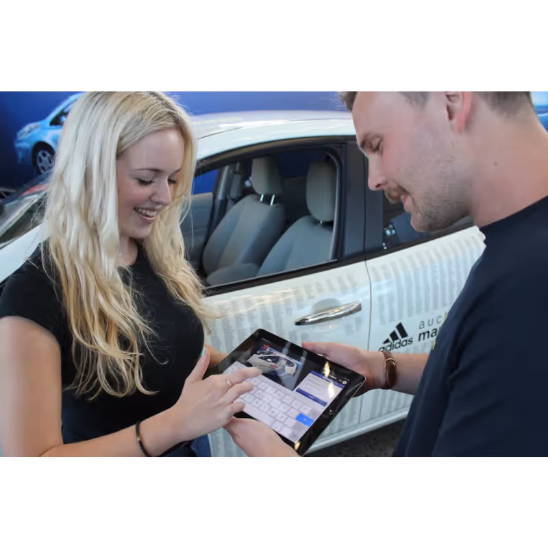 Woman and man using a tablet in front of a white car with Adidas and Auckland logos.