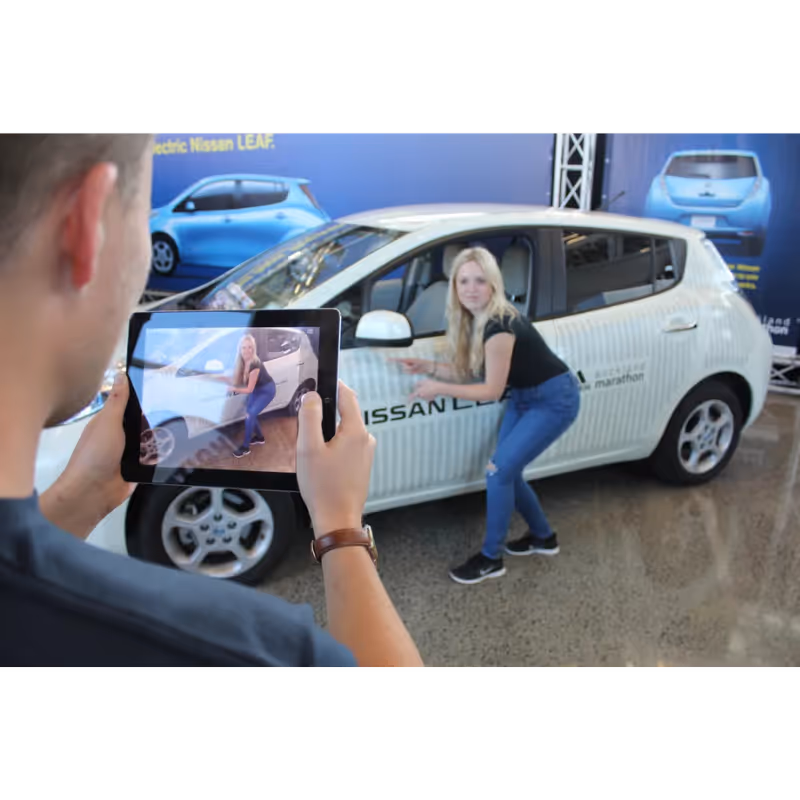 Man uses tablet to photograph woman posing beside white Nissan LEAF car indoors.