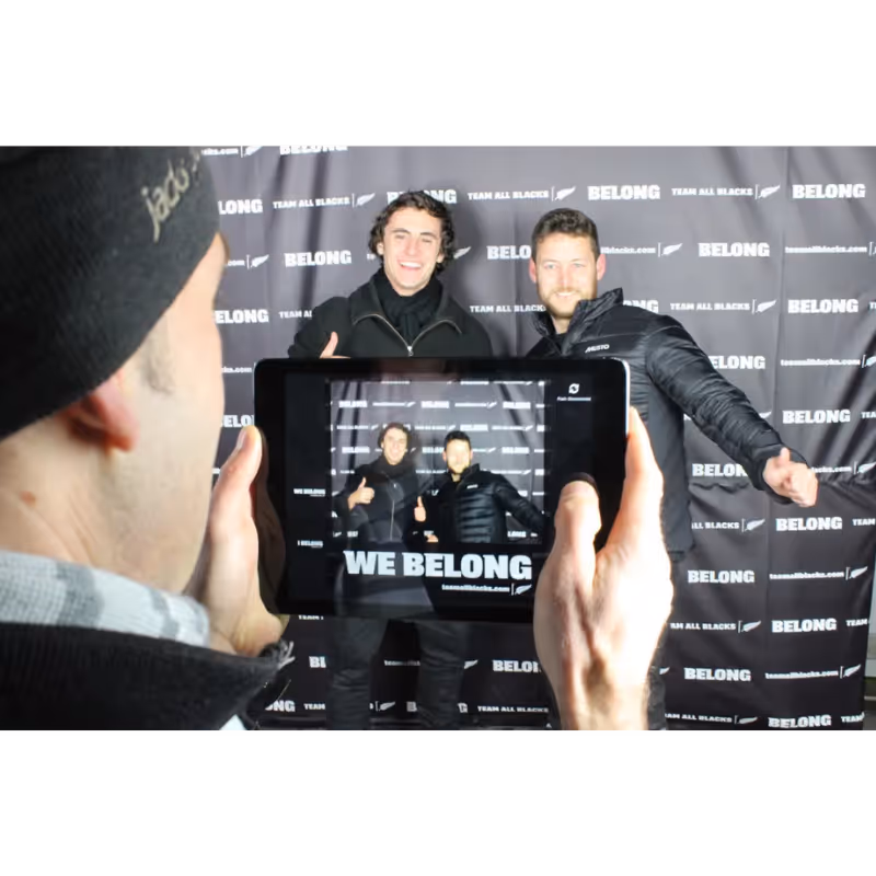 Man holding tablet showing two men posing with thumbs up in front of 'WE BELONG' backdrop.