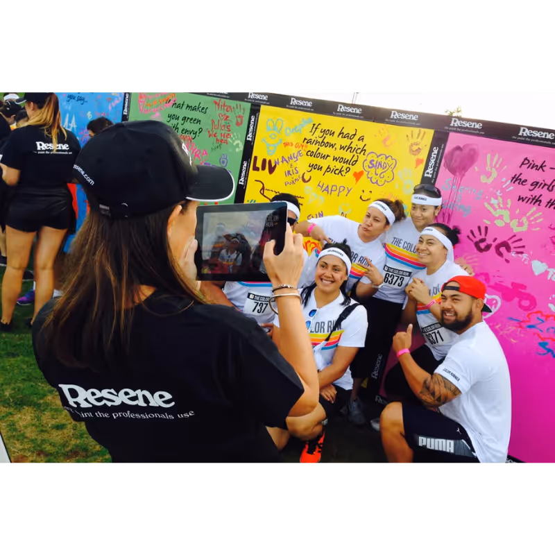 Woman in Resene shirt uses tablet to photograph smiling group posing in front of colorful walls at event.