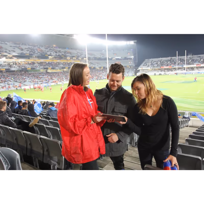 Three people standing in stadium seats, one holding a tablet, watching a nighttime sports game.