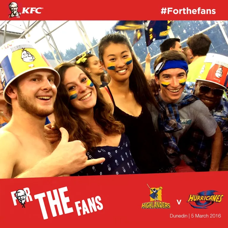 Group of smiling sports fans with blue and yellow face paint and KFC bucket hats at a Highlanders vs Hurricanes game.