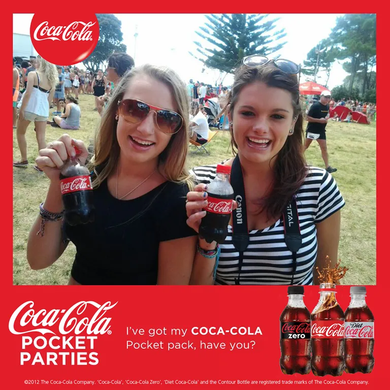 Two women smiling outdoors at event, holding Coca-Cola Pocket pack bottles with logo and product display.