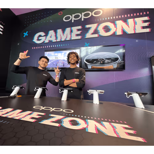 Two men smiling and posing at OPPO Game Zone booth with gaming controllers on the table.