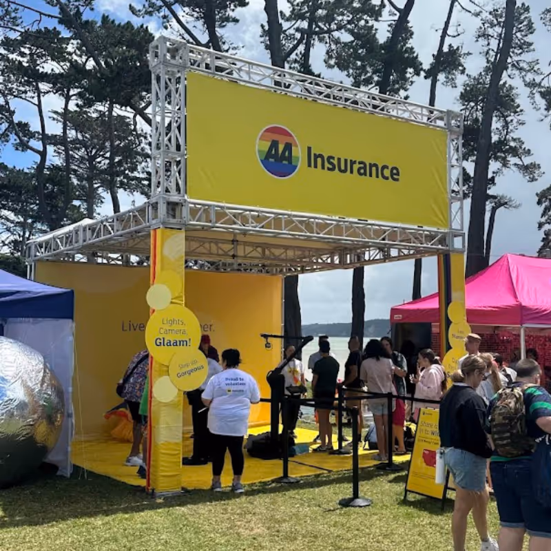 People gathered at a bright yellow A4 Insurance outdoor booth with trees and blue sky in the background.
