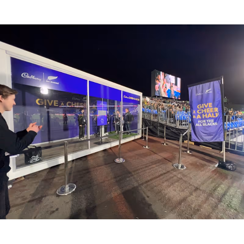 People clapping near a Cadbury and All Blacks cheer booth at a stadium event.