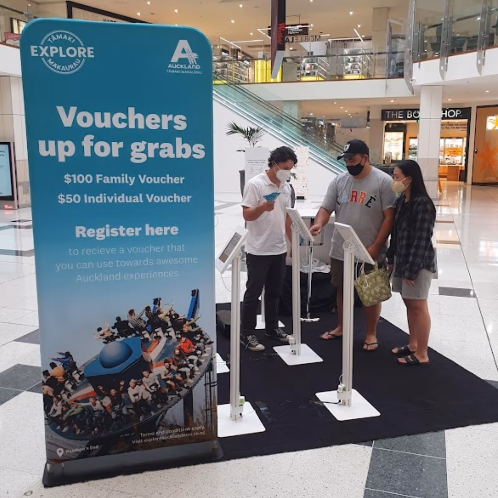 Three masked people registering for $50 and $100 Auckland vouchers at kiosks in a shopping mall.
