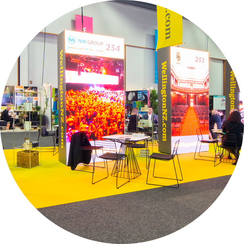 Bright exhibition booth with digital displays, chairs, and tables on a yellow carpet at an event.