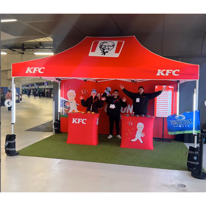 Three people waving under a red KFC tent with branded counters inside a covered area.