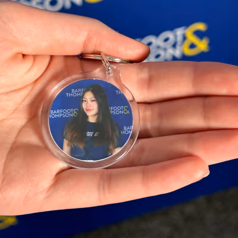 Hand holding a round keychain with a photo of a woman against a blue Barfoot & Thompson background.