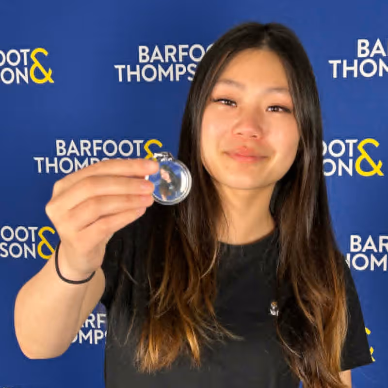 Woman with long dark hair holding a keychain in front of a Barfoot & Thompson backdrop.