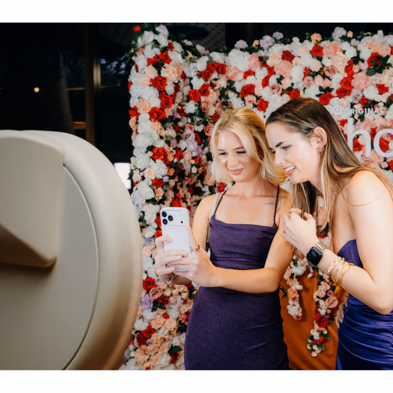 Two women in purple dresses smiling and taking a selfie with a phone in front of a flower wall.