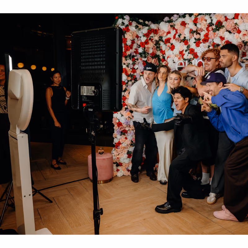 Group of seven people posing happily for a photo in front of a flower wall at an event.