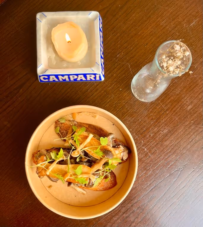 A dish by Bia, on a wooden table next to a glass vase and a vintage Campari ashtray with a lit candle sitting inside it