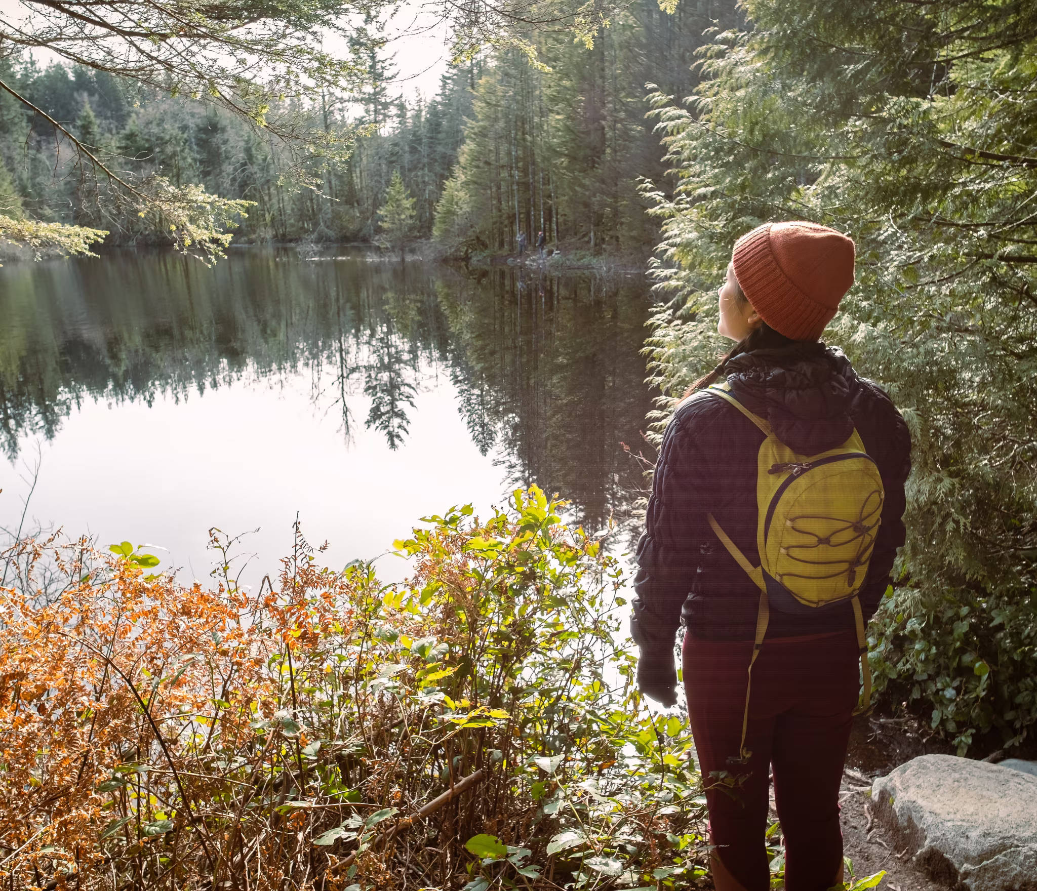 A person wearing a red beanie, dark jacket, and yellow backpack stands at the edge of a calm forest lake, looking out at the water surrounded by evergreen trees. Sunlight filters through the trees, illuminating the foliage and water’s surface.