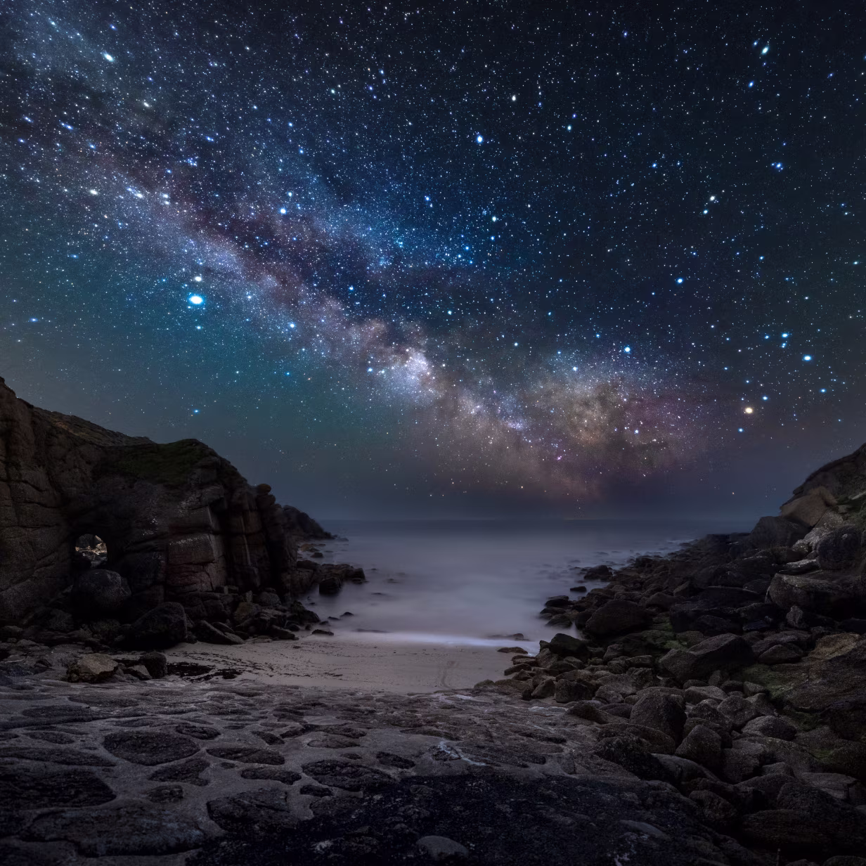A rocky shoreline under a star-filled night sky with the Milky Way stretching diagonally overhead. Soft waves lap against the beach, illuminated faintly by starlight.