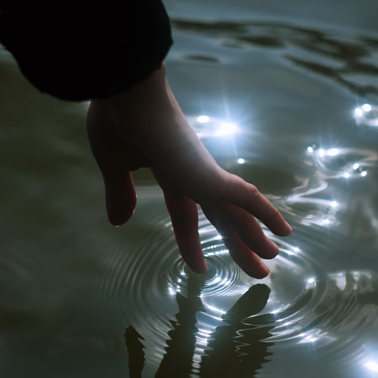 A hand gently touches the surface of calm water, creating ripples and reflections. Points of bright light sparkle on the water, contrasting with the shadows.